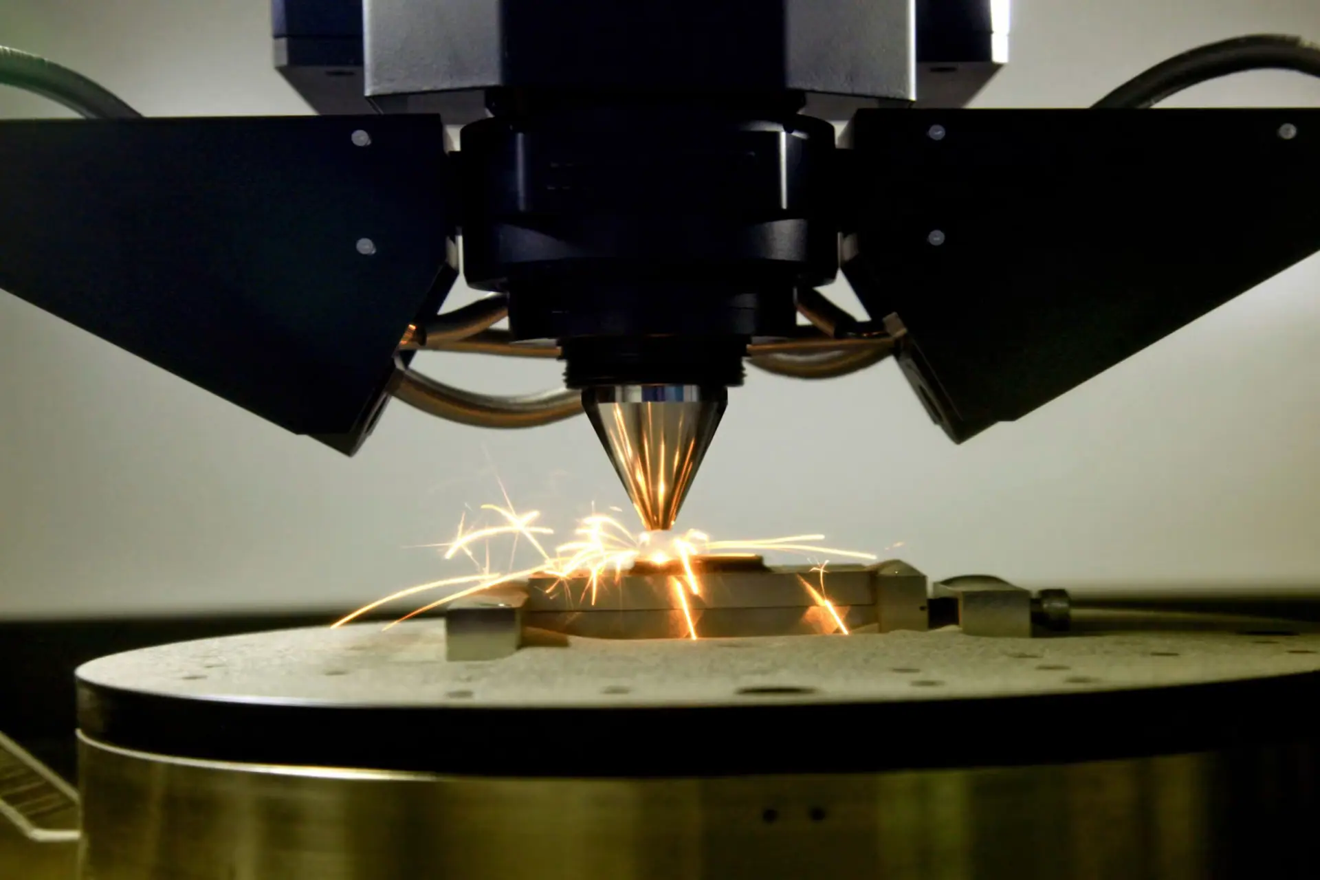 Laser melting metal powder on a build plate, sparks rising from the melt pool.